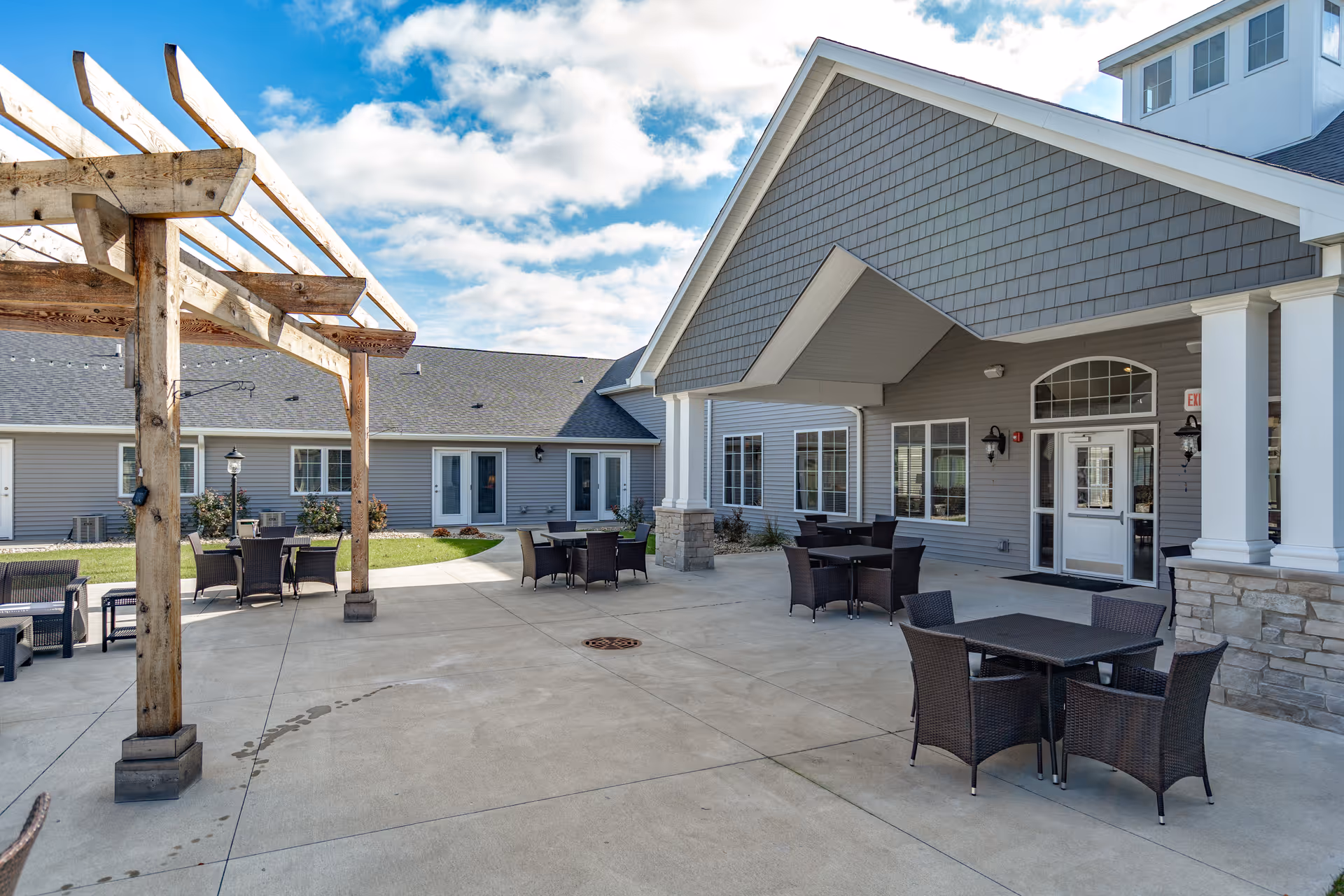 Outdoor patio area at Villas of Holly Brook Assisted Living & Memory Care in Pekin, IL, featuring several black wicker tables and chairs on a concrete surface, a wooden pergola structure on the left, and the building exterior with gray siding and white trim under a partly cloudy sky.