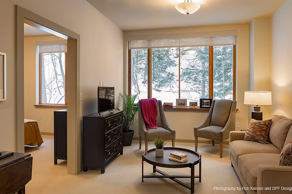 A cozy living room with a large window showing a snowy outdoor scene. The room features two gray armchairs, a beige sofa with patterned pillows, a round coffee table with books and a small plant, a black dresser with a TV on top, a floor lamp, and a potted plant near the window. An adjacent bedroom is partially visible through an open doorway.