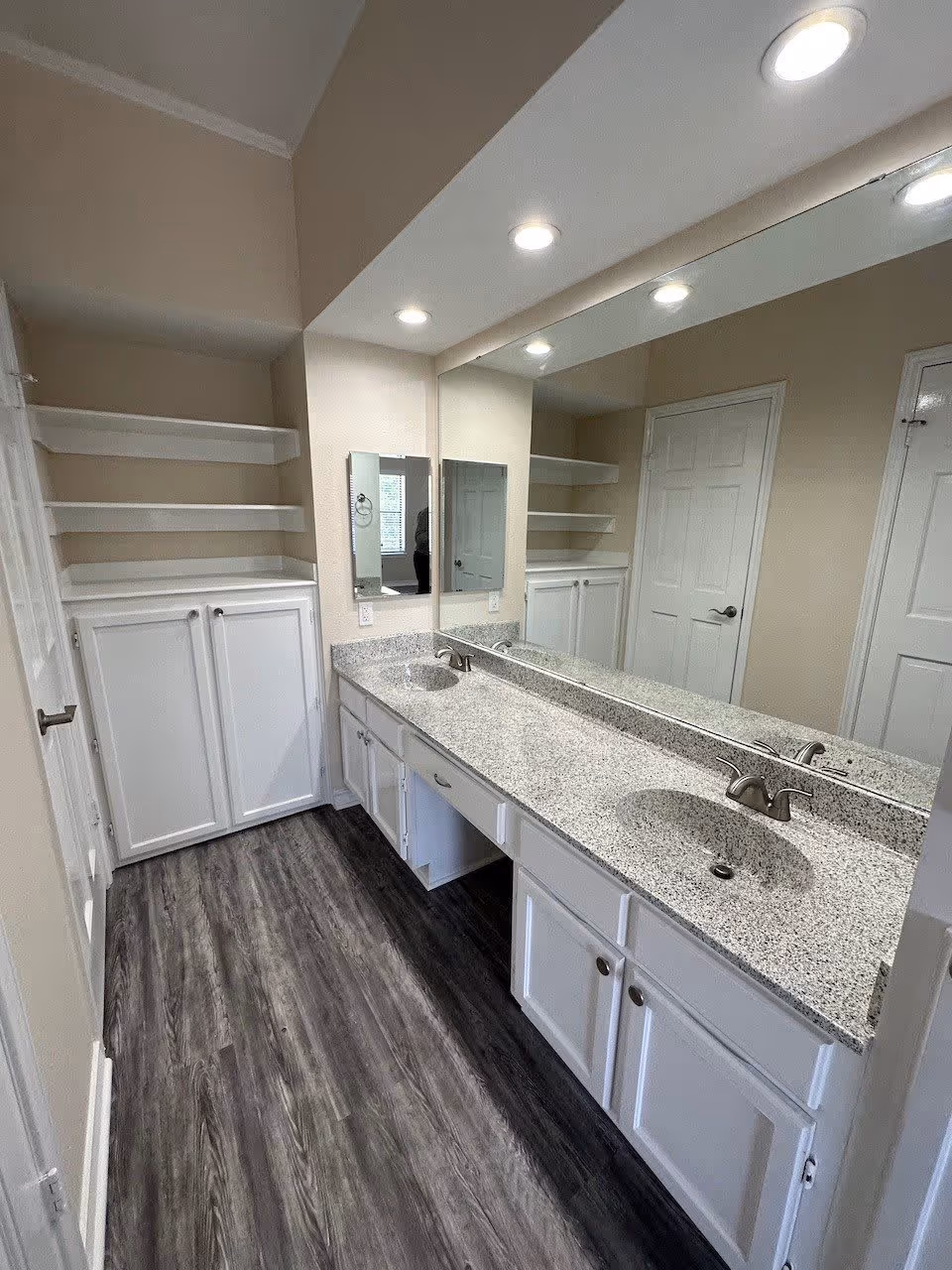 Long bathroom vanity with double sinks, a large mirror, white cabinets and open shelving under recessed ceiling lights.