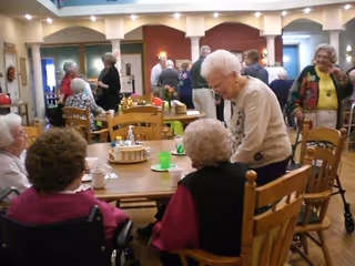 A group of elderly people socializing in a communal dining area with wooden tables and chairs. Some individuals are seated while others are standing and engaging in conversation. The room has warm lighting and a welcoming atmosphere.
