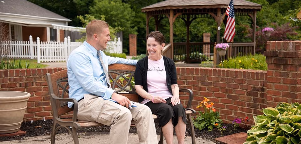 A man and an elderly woman sitting on a wooden bench in a garden area with brick walls, flowers, and a gazebo in the background. The man is wearing a light blue shirt and beige pants, while the woman is wearing a black cardigan and a light pink shirt. They are engaged in a friendly conversation.