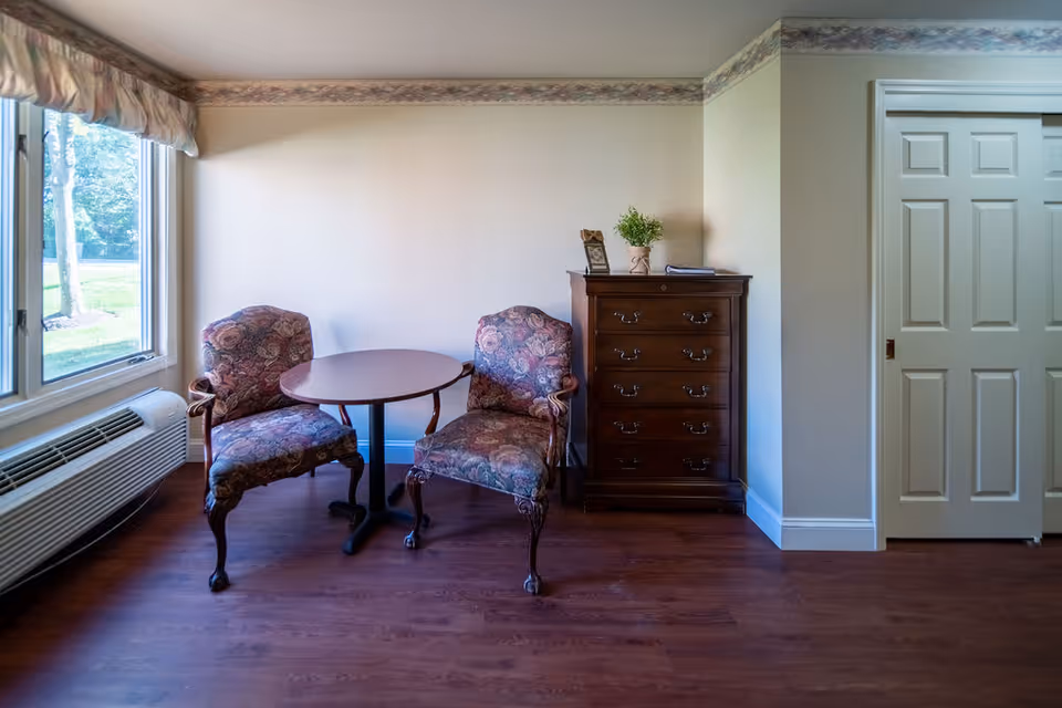 A small sitting area with two floral upholstered armchairs and a round wooden table between them. To the right is a dark wooden chest of drawers with a small plant and a framed photo on top. There is a large window on the left letting in natural light, and a white sliding door on the right side of the image. The floor is wooden and the walls are light-colored with a decorative border near the ceiling.