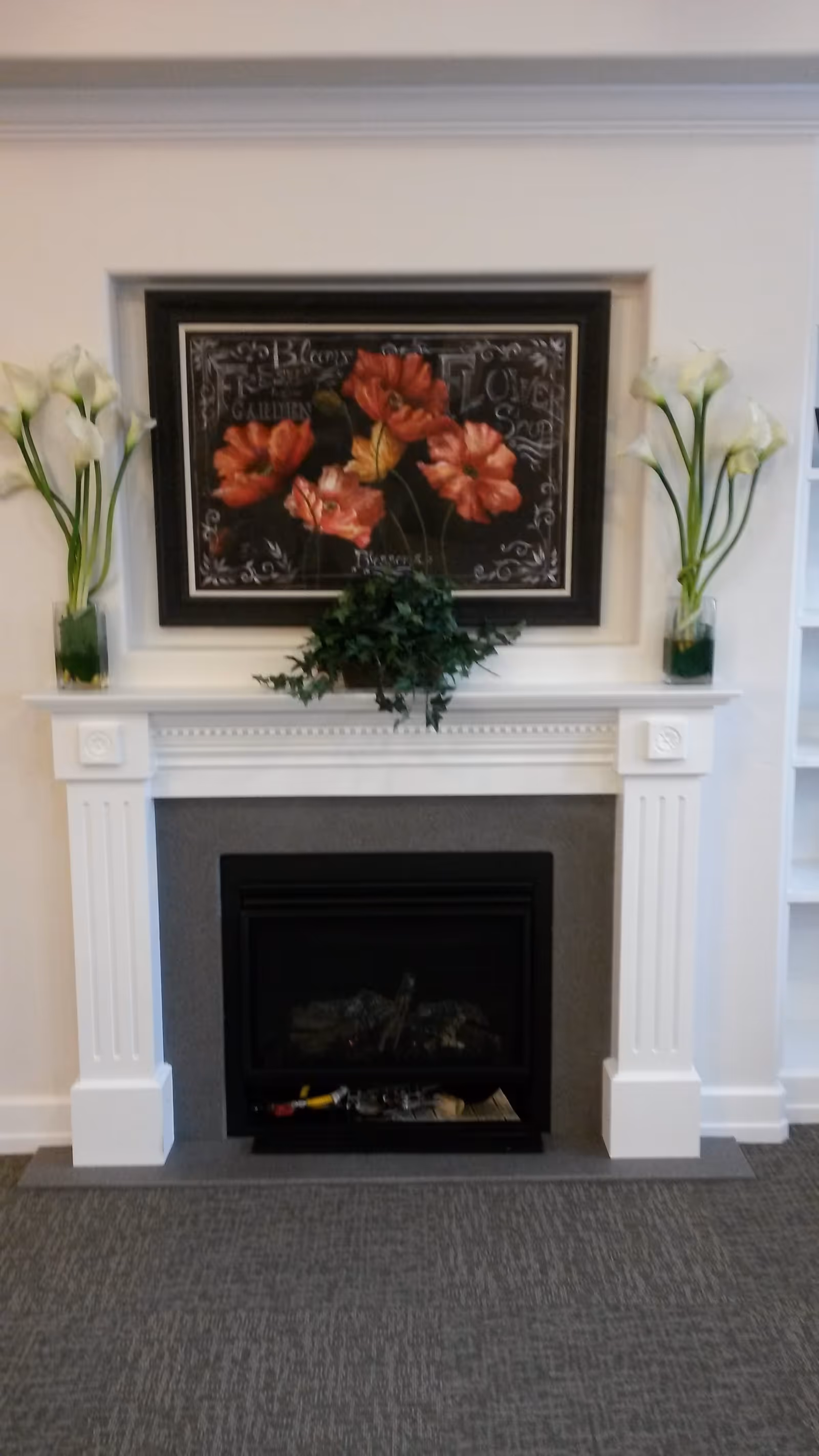 A white decorative fireplace with a black insert, flanked by two vases each holding white calla lilies. Above the fireplace is a framed artwork featuring red and orange flowers with a dark background. A green leafy plant is placed on the mantel.