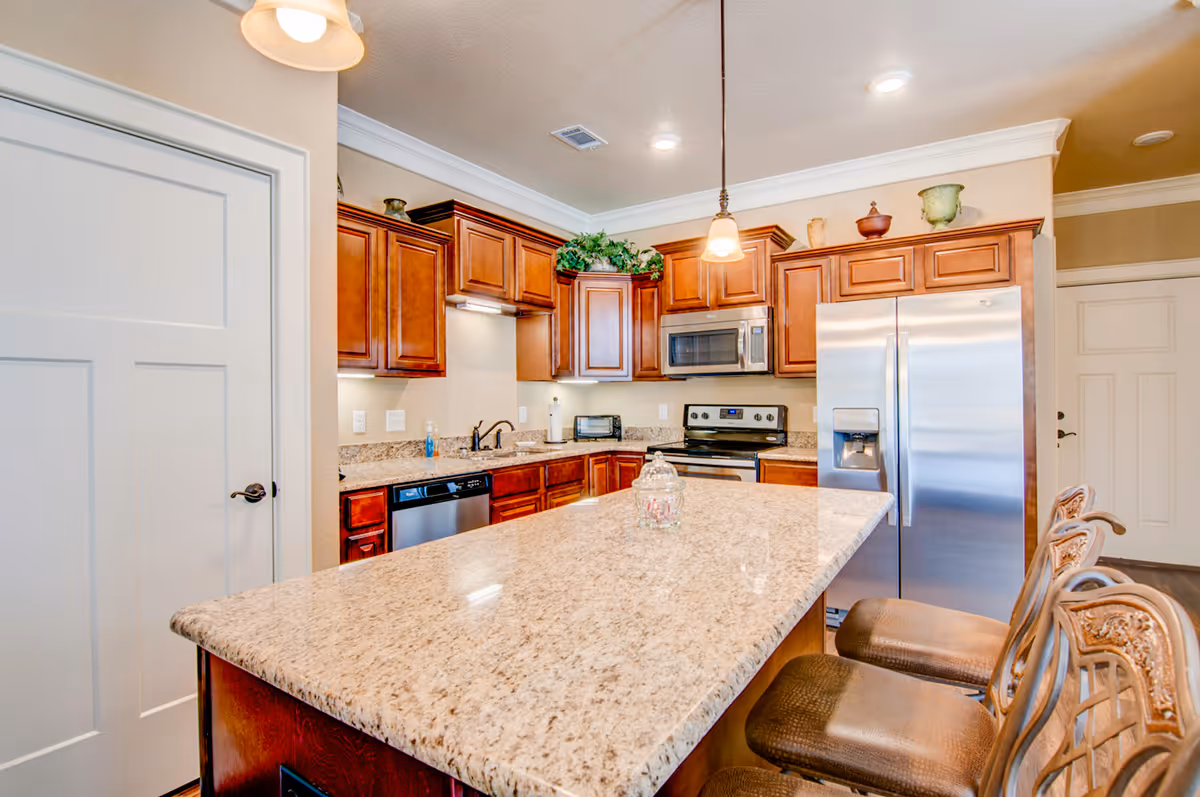 Bright modern kitchen with a large granite island, wooden cabinets, stainless steel appliances, and three bar stools.