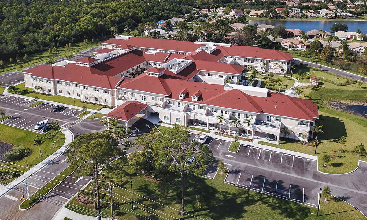 Aerial view of Alamar Senior Living facility showing a large building with red roofs surrounded by parking lots, green lawns, trees, and nearby residential houses with a water body in the background.