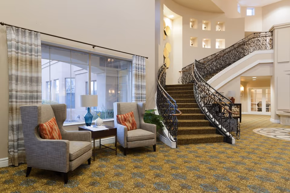 A spacious interior area featuring a curved staircase with ornate black railings and carpeted steps. Two gray upholstered armchairs with red patterned cushions are positioned near a window with striped curtains, separated by a small table holding a blue glass lamp and decorative items. The floor is covered with a patterned carpet, and the walls are painted in a light neutral color with small recessed display niches above the staircase.