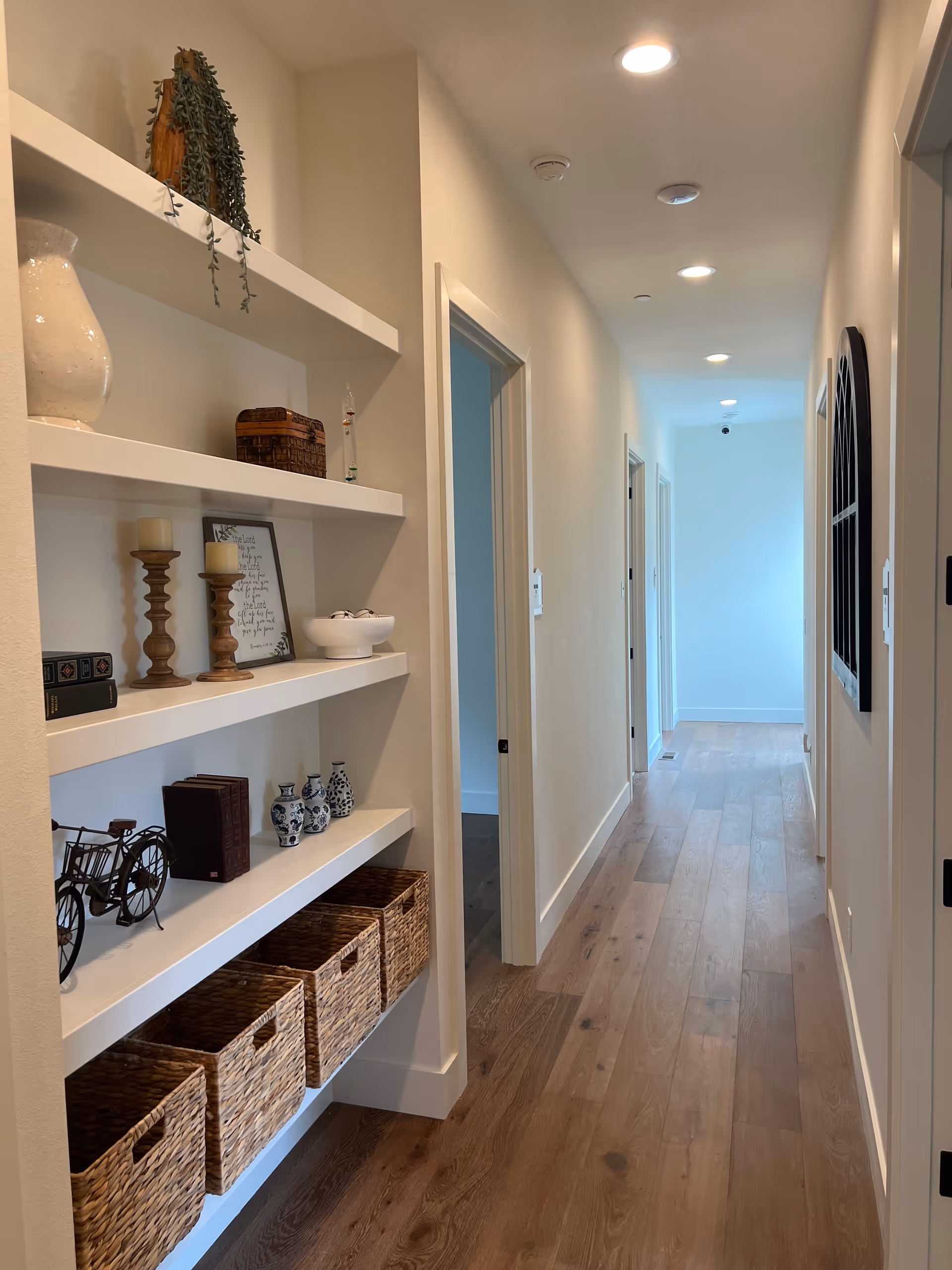 A well-lit hallway with wooden flooring and white walls. On the left side, there are built-in white shelves decorated with various items including candles, small vases, books, a small bicycle model, and woven baskets on the bottom shelf. Several doorways are visible along the hallway, and recessed ceiling lights illuminate the space.