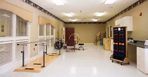 Bright rehabilitation room with parallel bars, exercise equipment, seating, and cabinets in a senior living facility.
