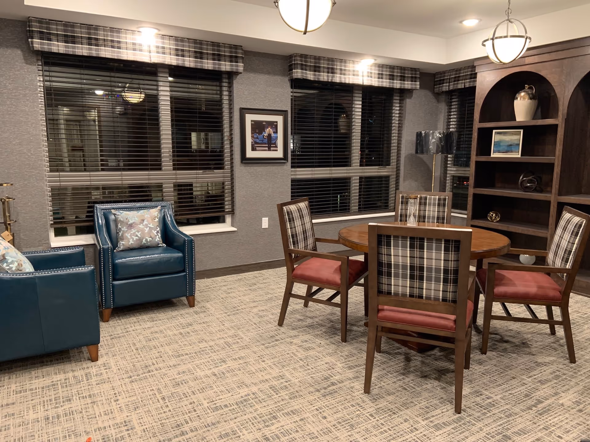 A cozy senior living common area with a round wooden table surrounded by four chairs with plaid backs and red seats. Two blue armchairs with decorative pillows are positioned near large windows with plaid valances and blinds. A dark wooden bookshelf with decorative items and a floor lamp are also visible. The room has patterned carpet and soft lighting.