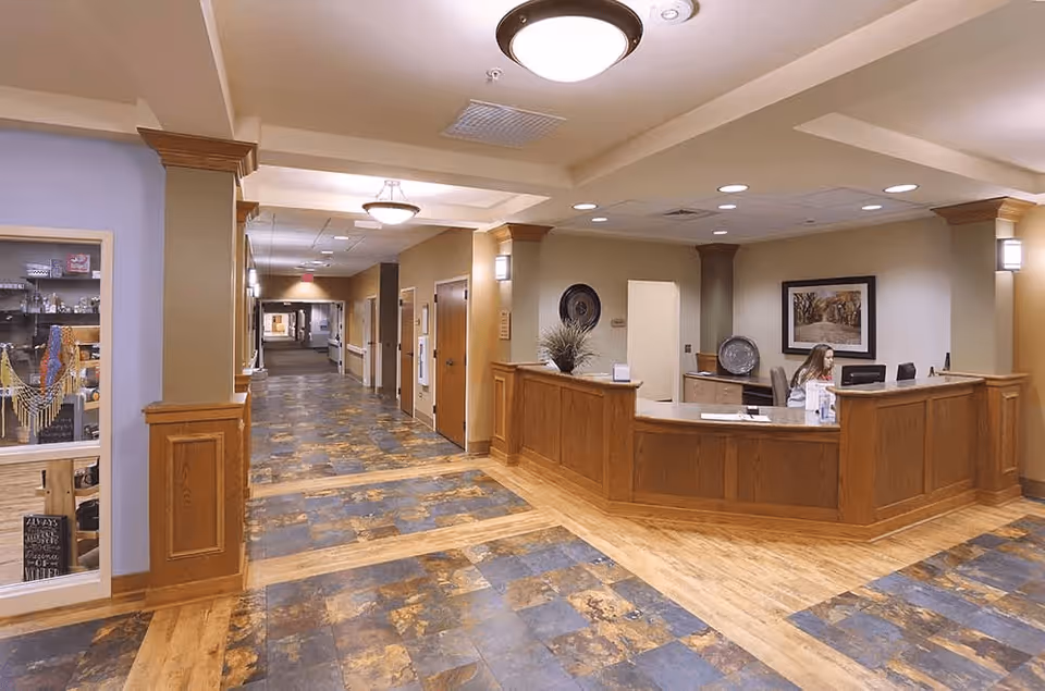 Interior view of a senior living facility reception area with a wooden front desk where a woman is seated working on a computer. The hallway extends into the distance with doors on either side and decorative lighting on the ceiling. The floor has a patterned tile and wood design, and there is a display case on the left side of the image.