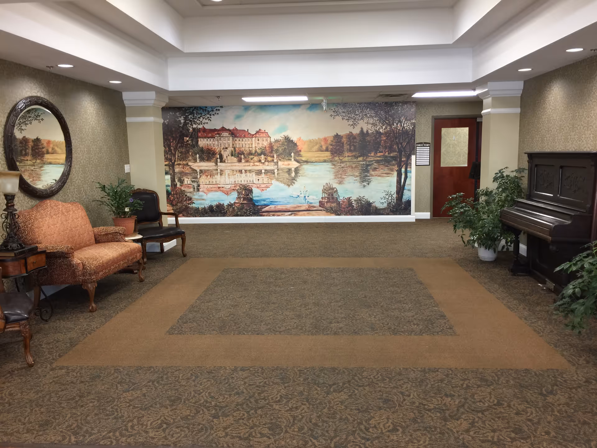 A carpeted lobby-like interior with upholstered seating, a piano, plants and a large lakeside estate mural on the far wall.