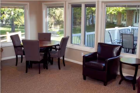 Sunlit lounge area with round table, chairs, leather armchair, and large windows.
