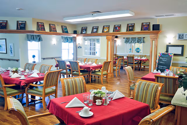 A dining room with several tables covered in red tablecloths, each set with white napkins, cups, glasses, and small flower arrangements. The room has wooden chairs with striped upholstery, large windows with curtains, and framed pictures displayed on a shelf near the ceiling. There is a chalkboard menu on the right side and a wooden floor.