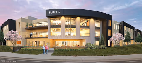 Exterior view of a modern three-story senior living facility named Solera, with large windows, balconies, and landscaped greenery including trees and bushes. Two people are walking on the sidewalk in front of the building under a partly cloudy sky.