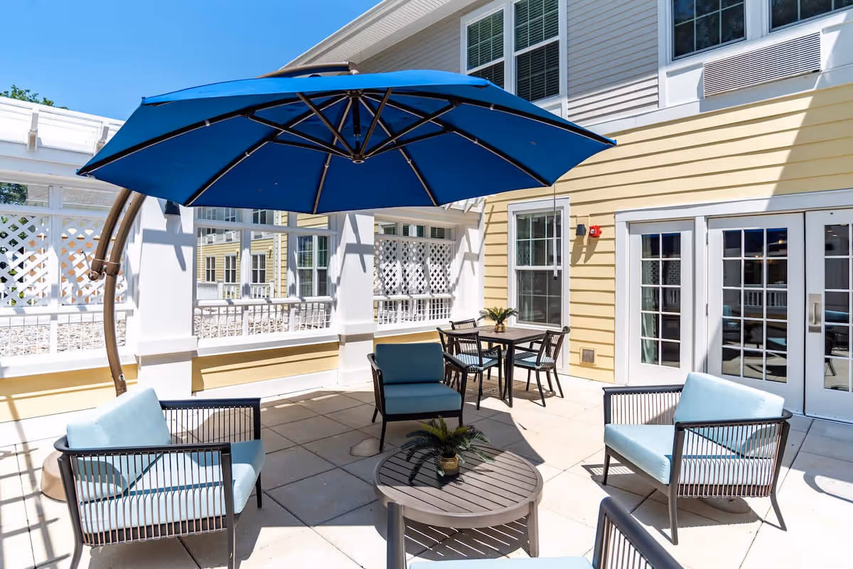 Outdoor patio area at Arbor Terrace Waugh Chapel with light blue cushioned chairs, a round table with a small plant, a dining table with four chairs, and a large blue umbrella providing shade. The patio is adjacent to a yellow building with white trim and multiple windows and glass doors.
