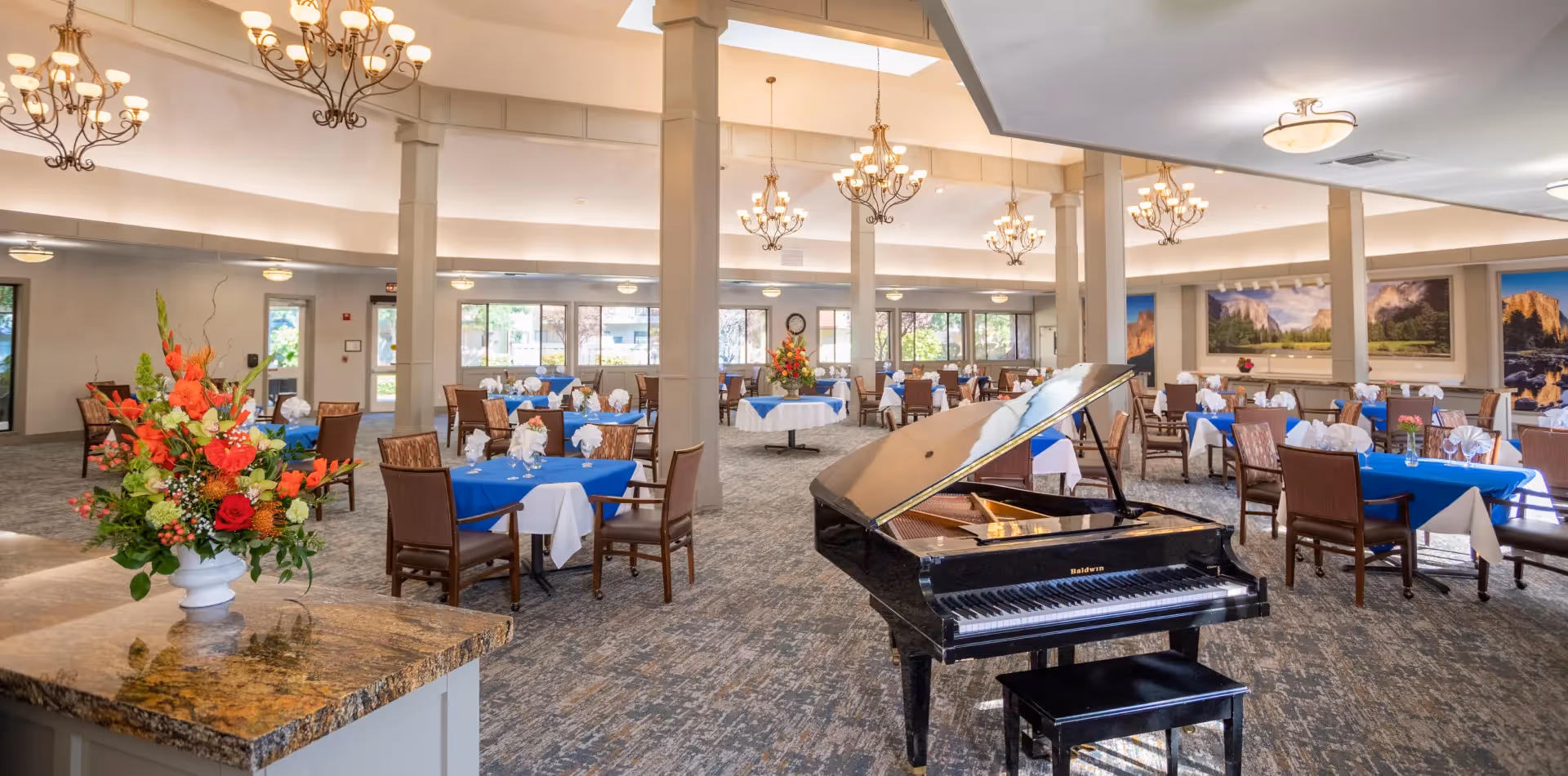 Spacious dining room with multiple tables covered in blue and white tablecloths, each set with napkins and glassware. A black grand piano is positioned in the foreground, and there are large floral arrangements on some tables and a countertop. The room is well-lit with chandeliers and large windows showing outdoor greenery. Landscape paintings adorn the walls.