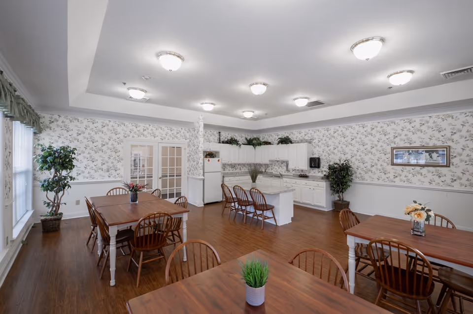 A bright dining room with wooden tables and chairs, floral wallpaper, and a kitchen area with white cabinets and a refrigerator. There are plants placed around the room and ceiling lights evenly spaced.