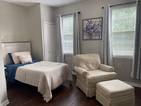 A bedroom with a single bed made with blue and beige bedding, a beige armchair with a matching ottoman, two windows with gray curtains, a closet with white doors, and a framed floral artwork on the wall.