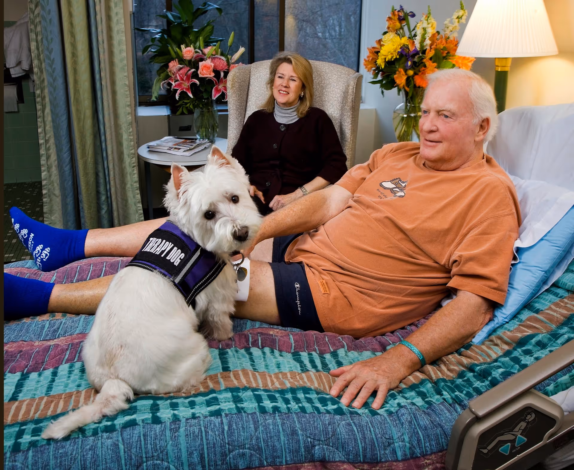 An elderly man is lying in a hospital bed wearing an orange t-shirt and blue socks, with a white therapy dog sitting on the bed near him. A woman is sitting in a chair behind the bed, smiling. The room has flowers on a table and a lamp in the background.