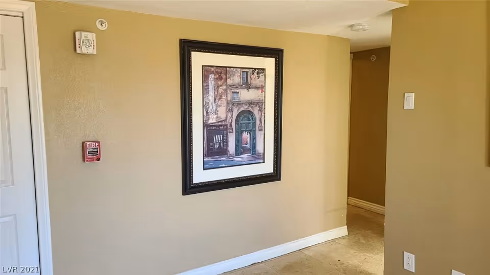 Interior hallway with beige walls and tiled floor. A framed picture of a building facade hangs on the wall. There is a fire alarm and a smoke detector mounted on the wall near a white door.
