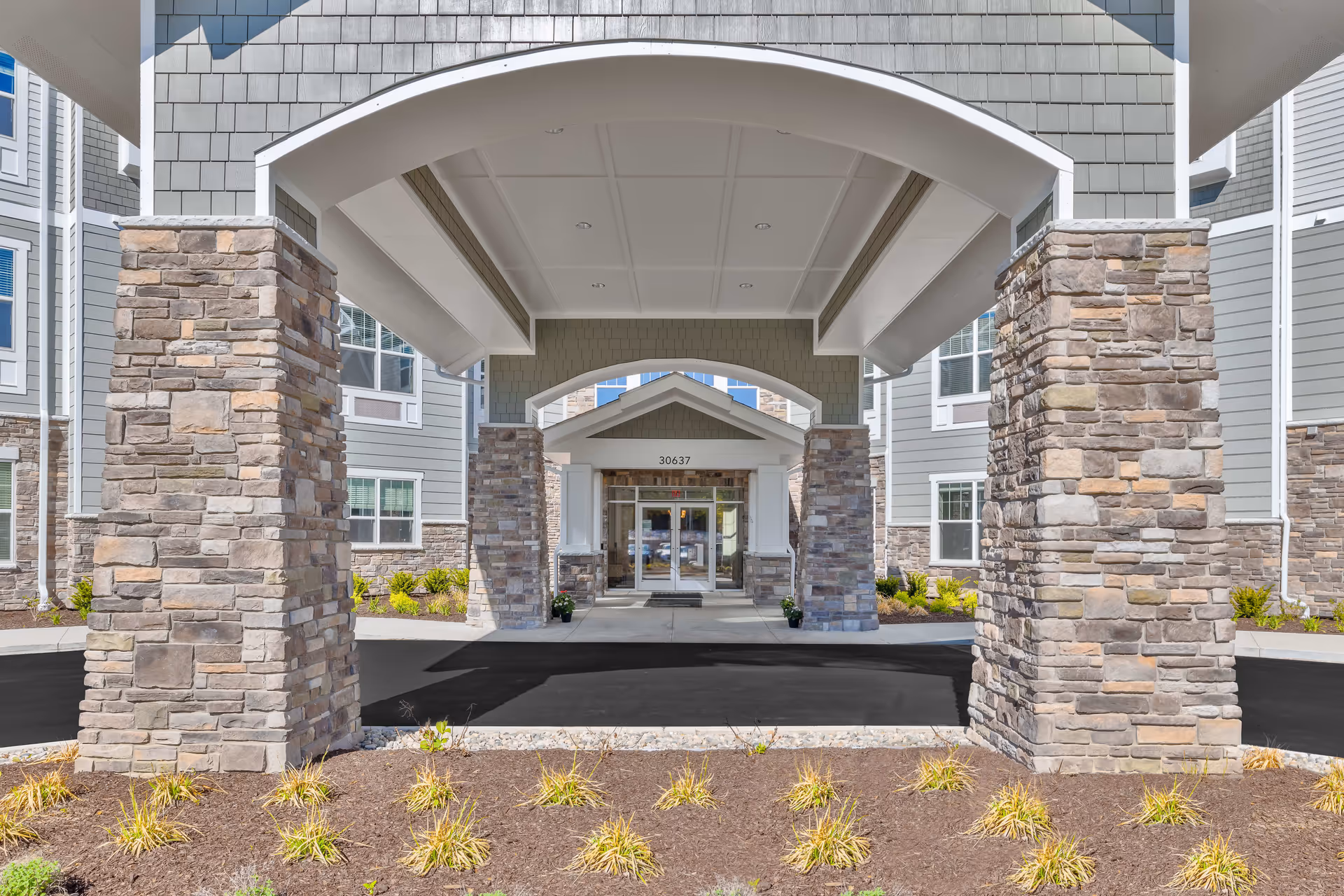 Covered entrance to a senior living facility with stone pillars and a driveway leading to glass double doors under a peaked roof. The building exterior features gray siding and multiple windows.
