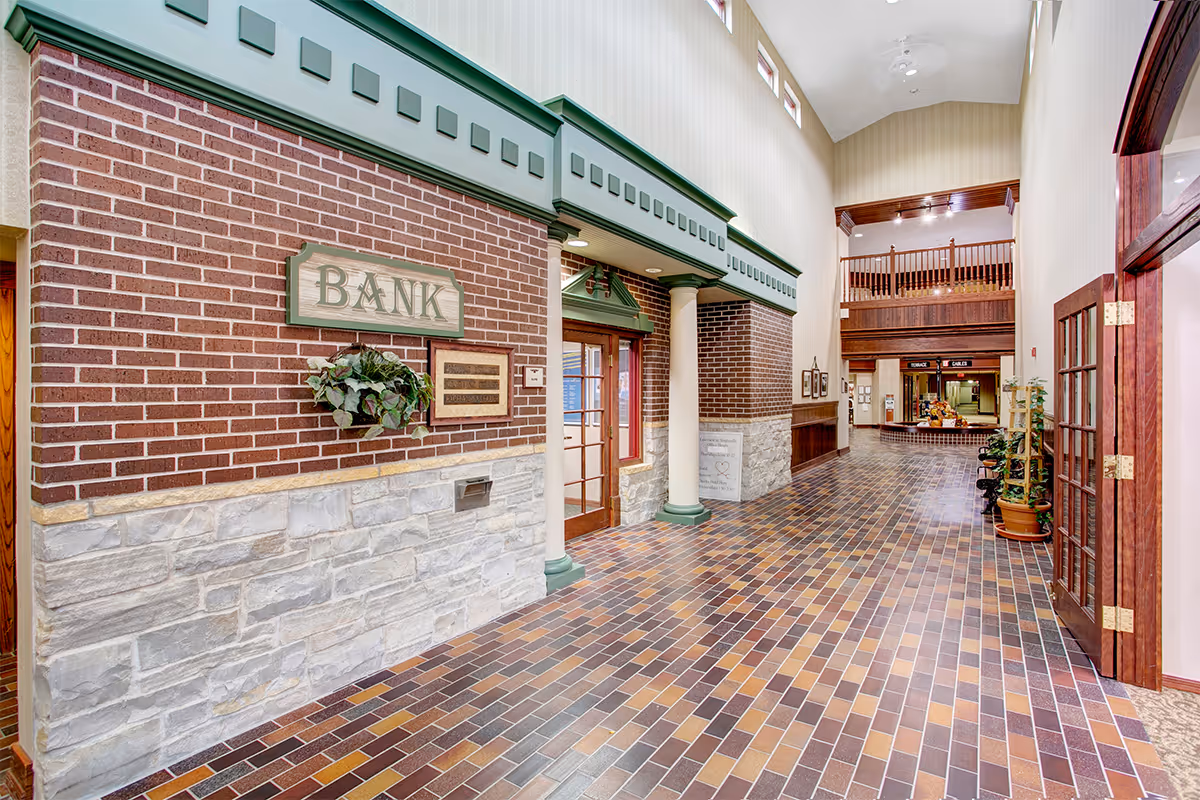 Interior hallway of a senior living facility with a faux bank facade on the left featuring brick and stone walls, green trim, and white columns. The floor is tiled with brown, yellow, and gray bricks. The hallway has high ceilings with small windows near the top and wooden railings on the upper level. There are plants and framed pictures along the walls.