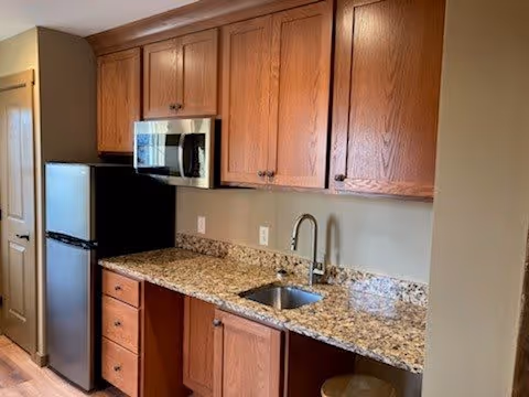 Interior view of a kitchen area featuring wooden cabinets, a granite countertop with a built-in sink and faucet, a microwave mounted above the counter, and a small refrigerator next to a closed door.
