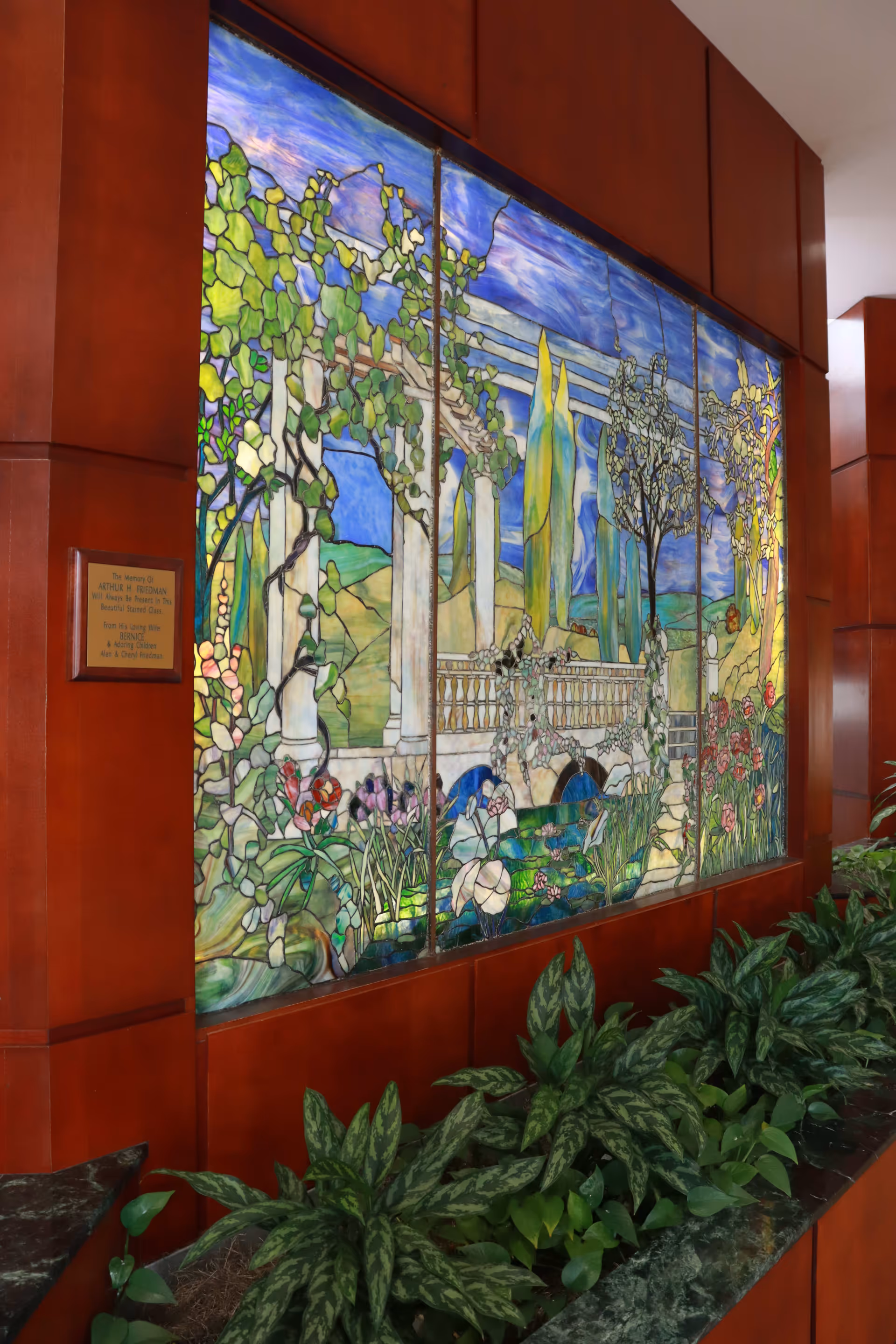 A large stained glass window depicting a scenic garden with trees, flowers, a stone bridge, and a blue sky, set within a wooden frame. Below the window is a planter filled with green leafy plants. A small plaque is mounted on the left side of the wooden frame.