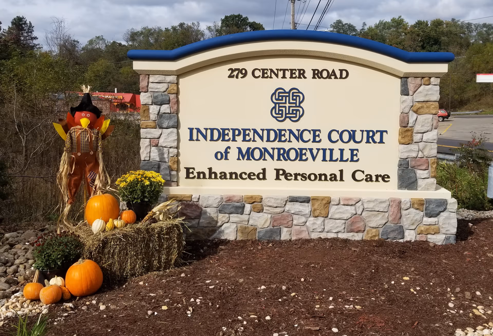 Outdoor stone sign reading Independence Court of Monroeville Enhanced Personal Care, autumn pumpkins nearby.