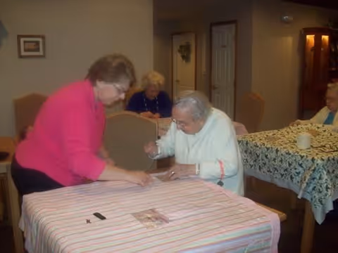 An elderly woman in a white sweater is seated at a table covered with a striped tablecloth, signing or writing on a piece of paper, while a woman in a pink top leans over to assist her. In the background, two other elderly women are seated at tables with floral tablecloths in a softly lit room.