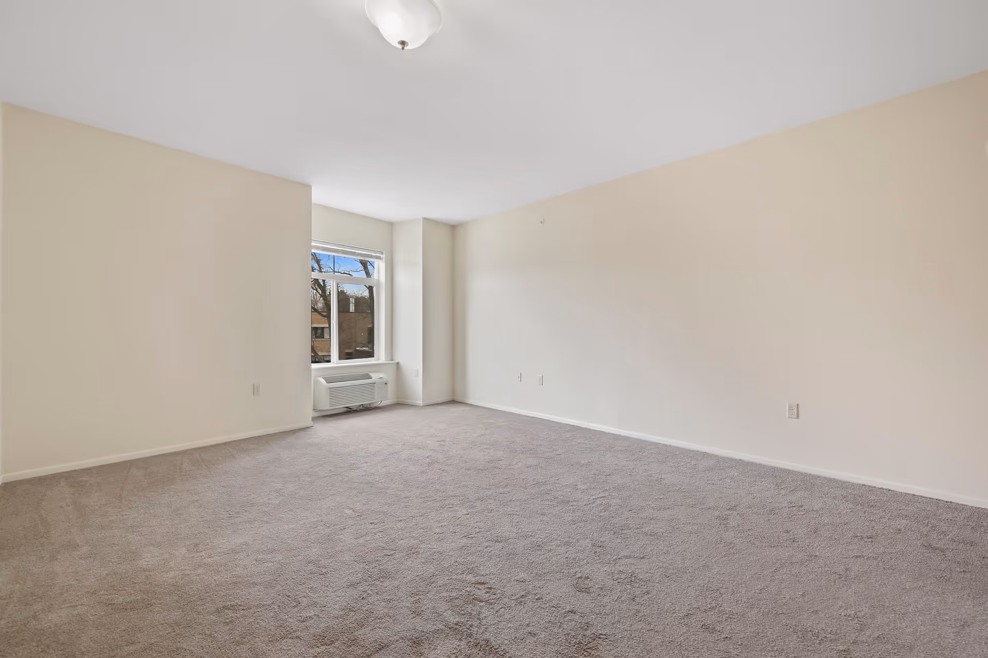 Empty room with beige walls and carpeted floor, a window with a view of trees and buildings outside, and a wall-mounted air conditioning unit below the window.