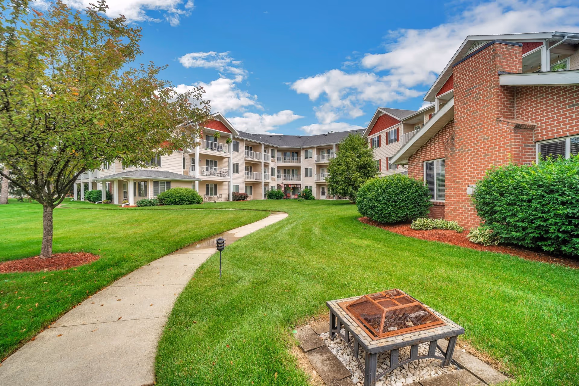 Outdoor view of a senior living facility with a curved concrete pathway, green lawn, trees, shrubs, and a fire pit in the foreground. The building has multiple floors with balconies and a mix of brick and siding exterior under a partly cloudy blue sky.