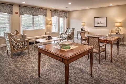 A well-lit living room area with large windows covered by patterned valances. The room features two beige sofas, two patterned armchairs, two wooden tables with chairs, and a floor lamp. The carpet has a subtle floral pattern, and there is framed artwork on the wall.