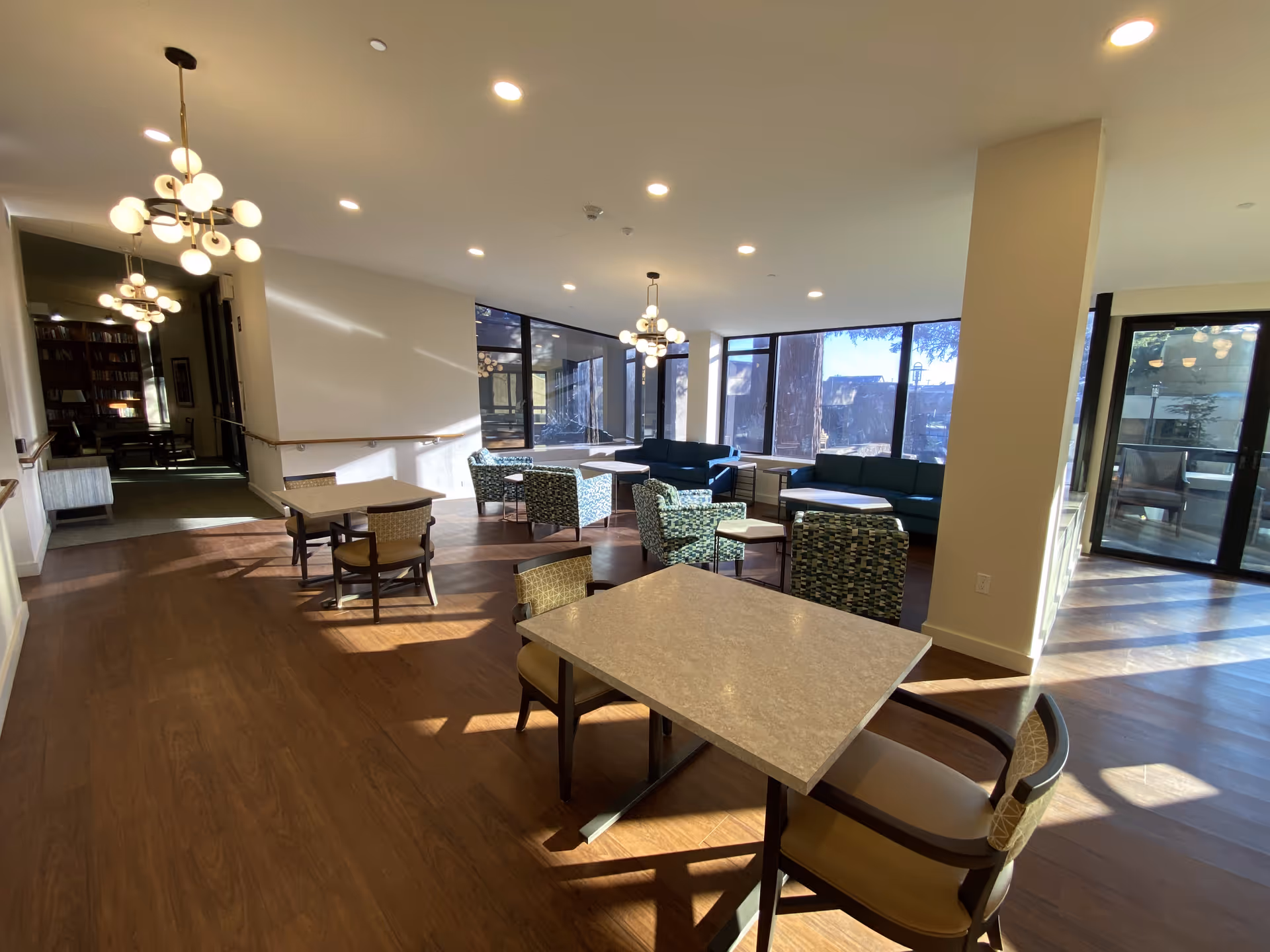 A bright and spacious common area in a senior living facility with multiple tables and chairs arranged on a wooden floor. The room features large windows allowing natural light to fill the space, modern pendant lights hanging from the ceiling, and comfortable seating areas with patterned armchairs and sofas. There is a doorway leading to another room with bookshelves visible in the background.