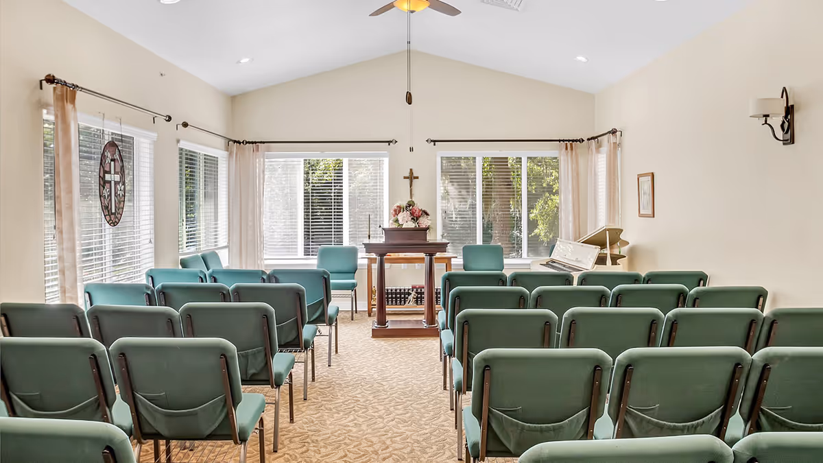 Interior view of a small chapel or meeting room with rows of green chairs facing a wooden podium adorned with a floral arrangement and a cross on the wall behind it. Large windows with blinds and light curtains let in natural light. A white piano is positioned to the right side of the podium.