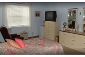 A bedroom with a bed covered in a floral bedspread and pink pillows. There is a brown recliner chair next to a window with closed blinds. Across from the bed is a dresser with a television on top and a larger dresser with a mirror above it. Various decorative items and framed pictures are visible on the walls and dressers.