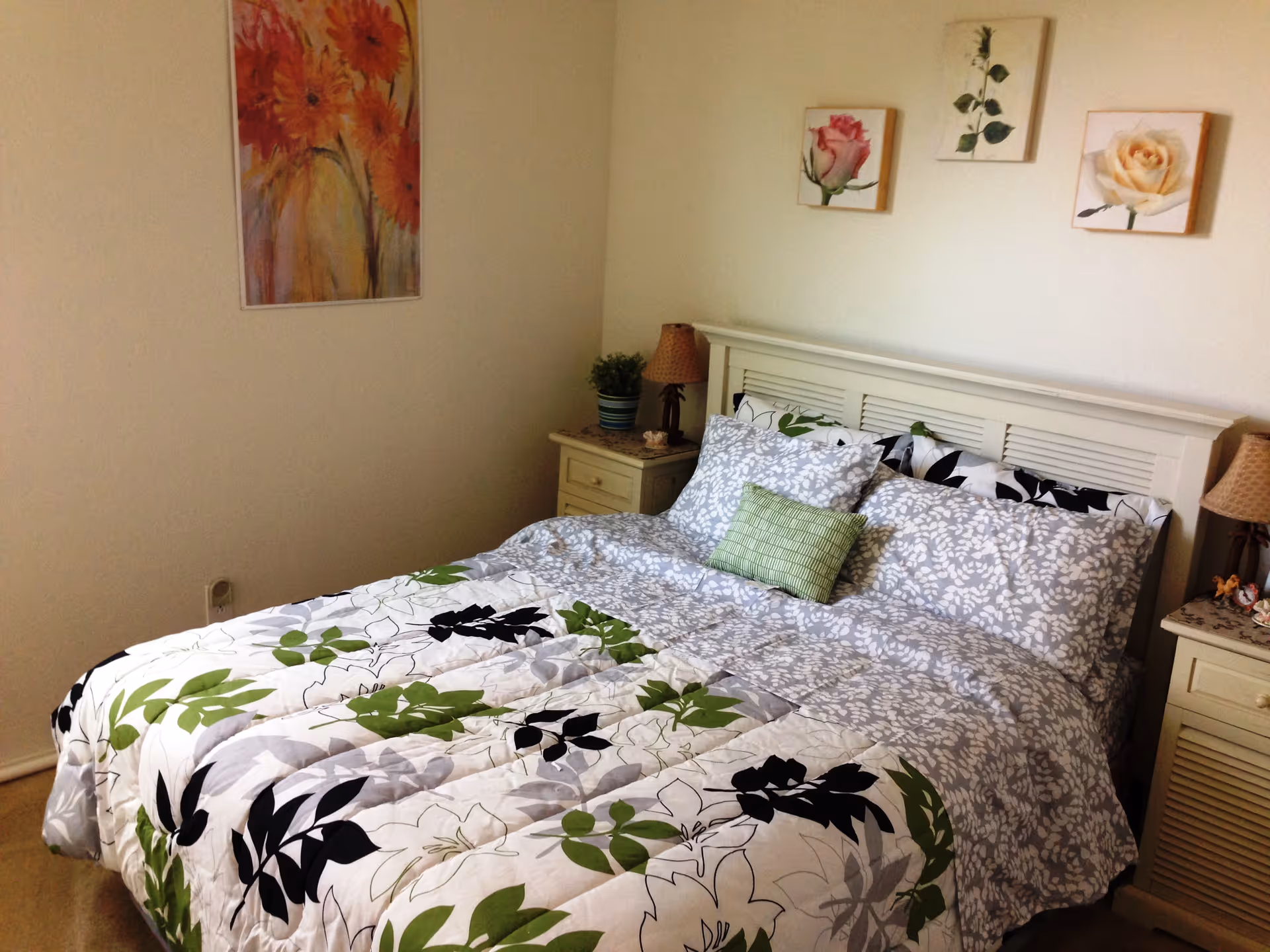 A neatly made bed with a white headboard and floral-patterned bedding in shades of green, black, and gray. There are two matching nightstands on either side of the bed, each with a lamp and decorative items. The walls are decorated with framed floral artwork.