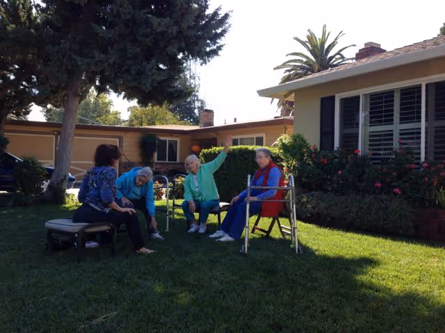 Four elderly women sitting and engaging in conversation on a grassy lawn outside a single-story house with bushes and trees in the background. One woman is seated on a bench, two women are seated on chairs, and one woman is sitting on the grass. The setting appears sunny and peaceful.