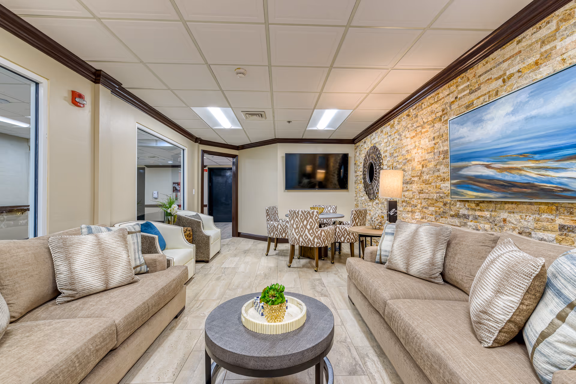 A cozy senior living common area with two beige sofas adorned with patterned pillows facing each other across a round gray coffee table with a decorative tray and plant. The room features a stone accent wall with a large landscape painting, a round mirror, and a table lamp. In the background, there is a round dining table with four patterned chairs and a wall-mounted flat-screen TV. The space has light wood flooring, beige walls, and a white tiled ceiling with recessed lighting.