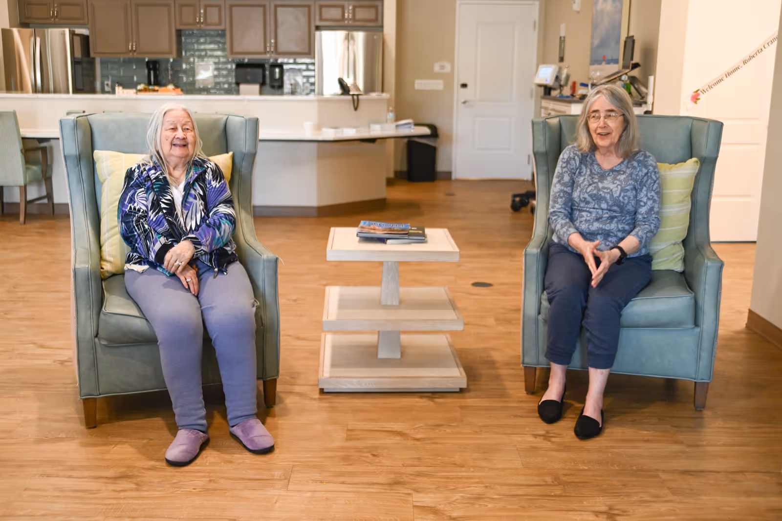 Two elderly women sitting in green armchairs with yellow pillows in a spacious room with wooden flooring. Between them is a small wooden table with books on it. In the background, there is a kitchen area with cabinets, a refrigerator, and a counter.