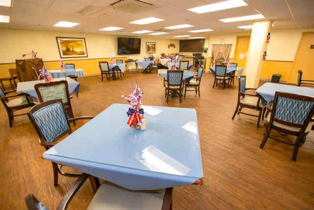 A spacious dining room with multiple tables covered in blue tablecloths, each decorated with small patriotic centerpieces. The room has wooden flooring, several chairs around each table, a large flat-screen TV mounted on the wall, and framed artwork. The walls are painted yellow and white, and the ceiling has recessed lighting.