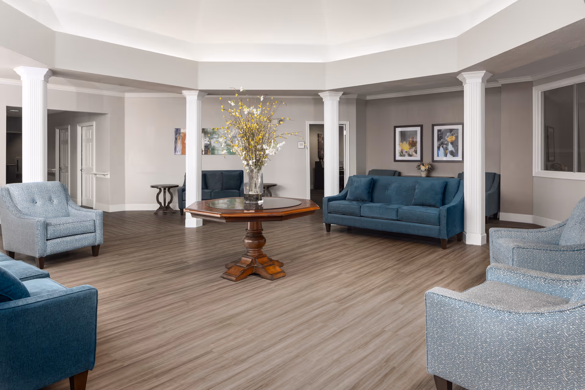 A spacious and well-lit living room area with light wood flooring, white columns, and a central wooden table with a vase of yellow and white flowers. The room is furnished with blue upholstered sofas and armchairs, and decorated with framed artwork on the walls.