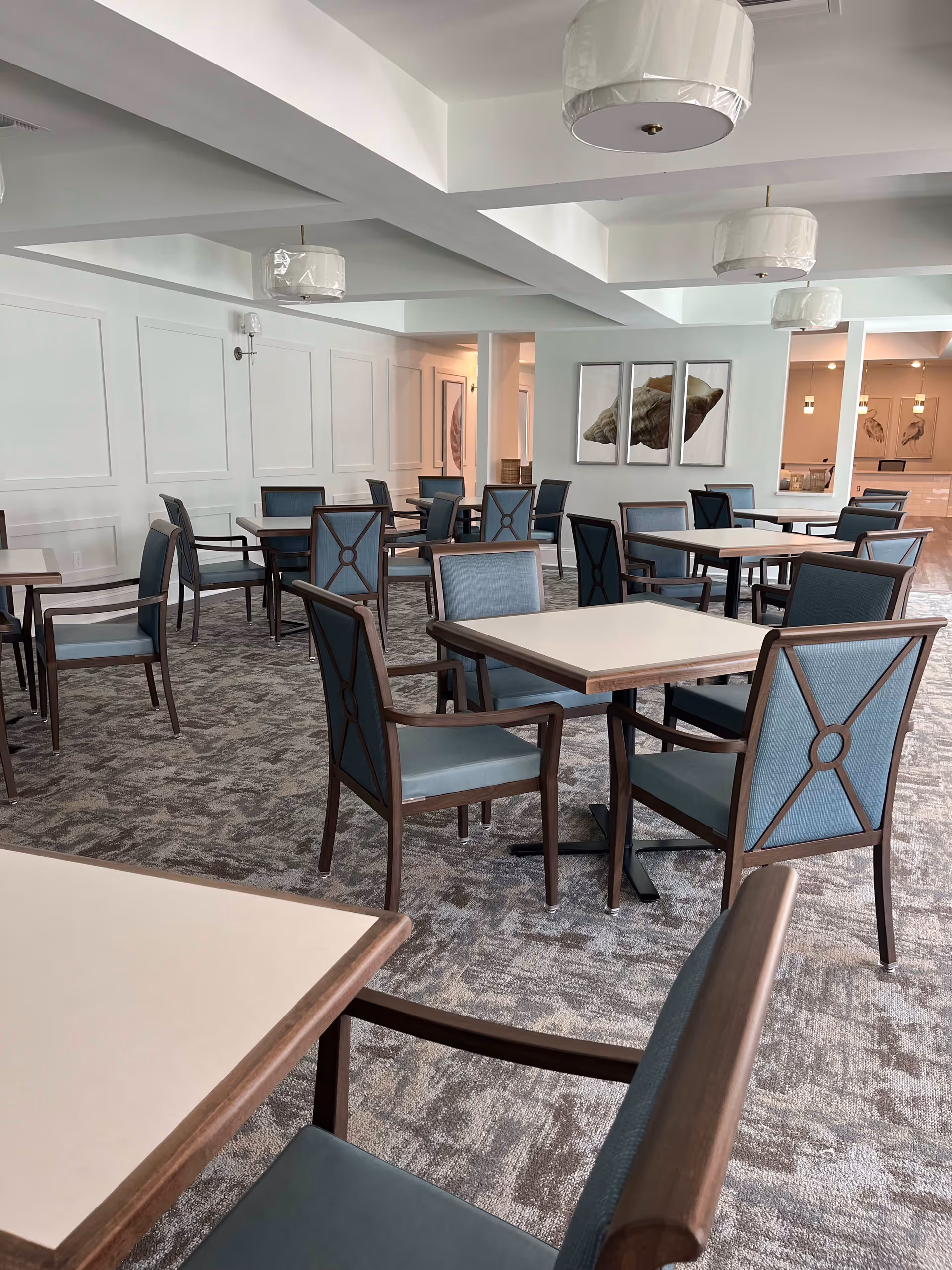 A spacious dining room with multiple square tables and blue cushioned chairs with wooden frames. The room has a patterned carpet, white walls with decorative paneling, and several modern ceiling light fixtures covered in protective plastic. There are framed artworks on the walls, including a large triptych featuring a seashell.