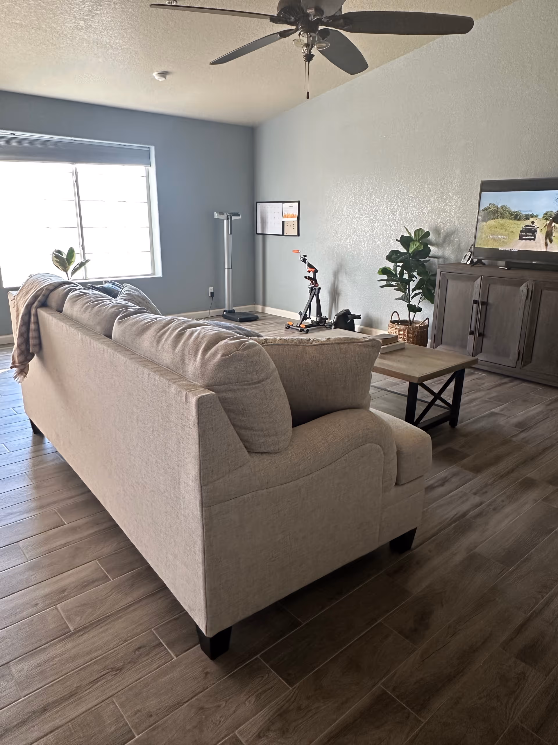 A living room with a beige couch facing a wooden TV stand with a flat-screen TV showing a nature scene. There is a ceiling fan above, a coffee table in front of the couch, a potted plant beside the TV stand, and exercise equipment including a scale and a scooter near the corner. The room has wood-look tile flooring and a window with blinds partially open letting in natural light.