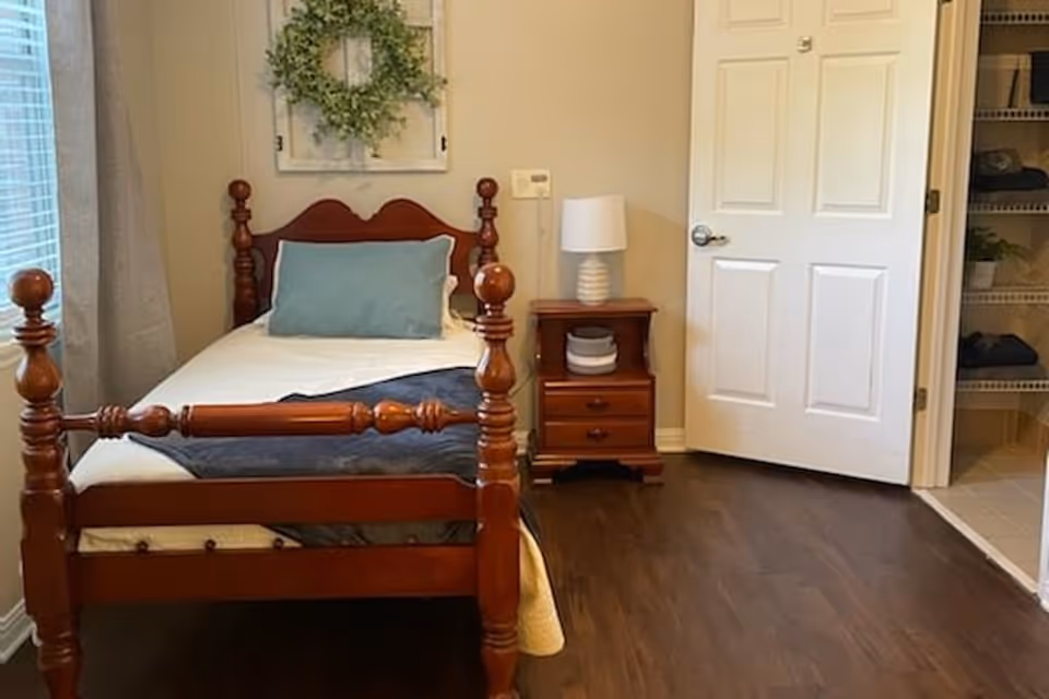 Small bedroom with a wooden single bed, bedside table and lamp, an open closet door, and wood flooring.