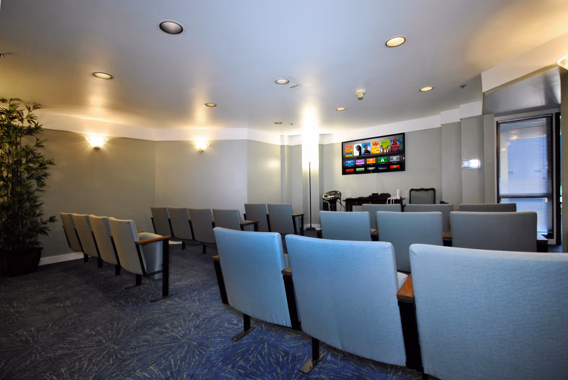 A small theater or presentation room with rows of cushioned chairs facing a wall-mounted flat screen TV. The room has soft lighting from wall sconces and recessed ceiling lights, a blue patterned carpet, and a potted plant in the corner. There is a door with blinds on the right side.