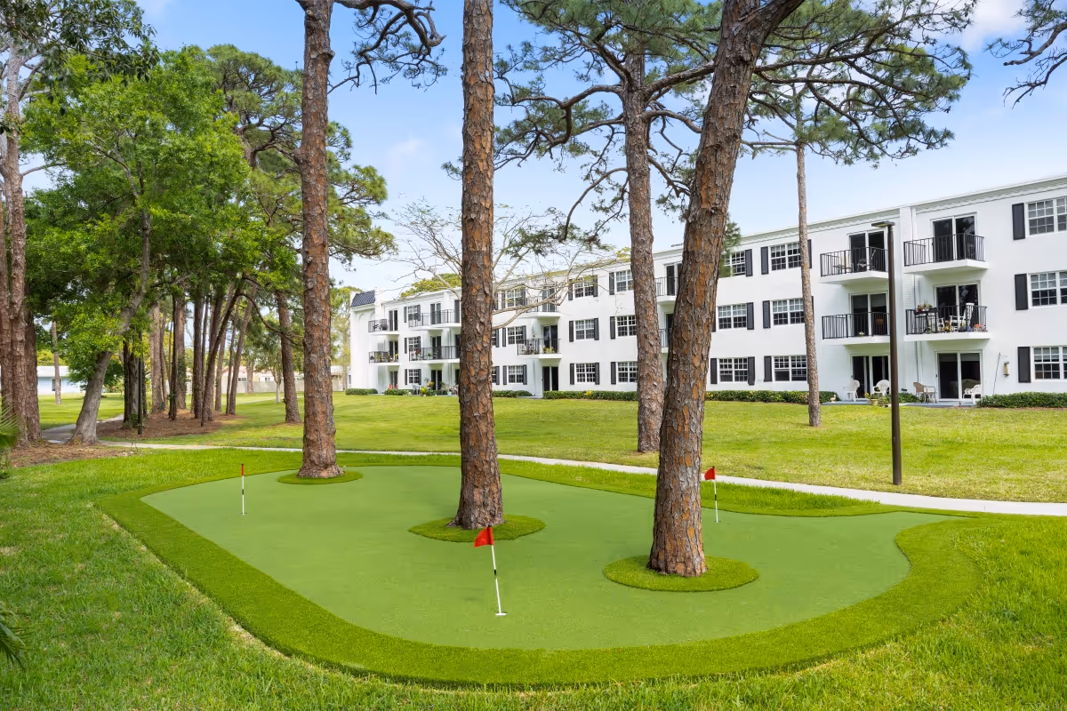 Outdoor putting green with three small red flags and several tall pine trees growing through the green, situated in front of a white three-story senior living building with balconies and black window shutters under a blue sky.