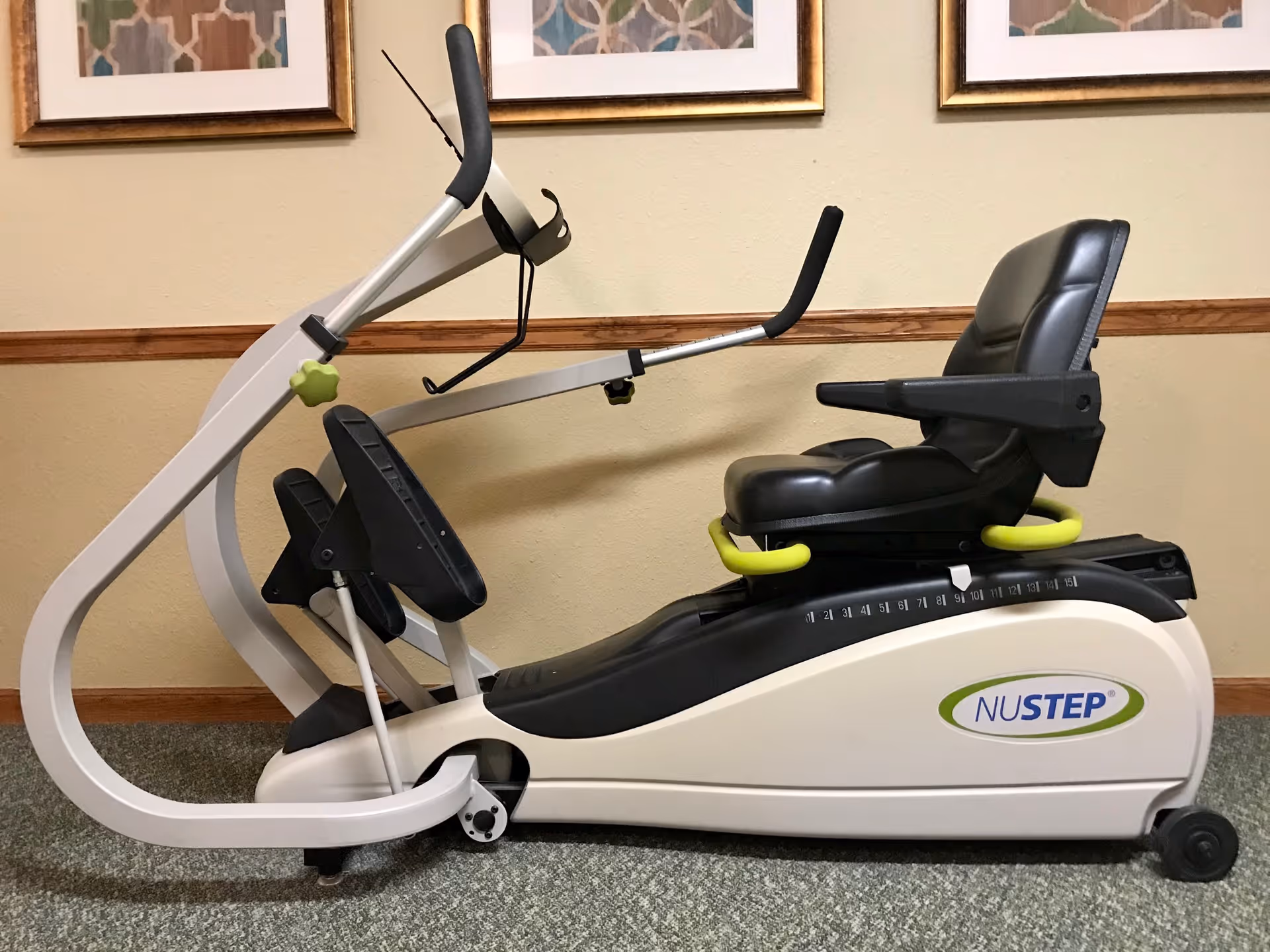 A NuStep recumbent cross trainer exercise machine with a black padded seat and arm handles, placed indoors against a beige wall with three framed abstract artworks hanging above it.