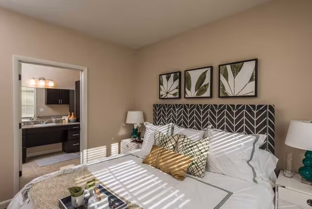 A well-lit bedroom with a large bed featuring a patterned headboard and multiple pillows, including a gold accent pillow. There are two bedside tables with matching green lamps. Three framed botanical prints hang above the bed. An open doorway reveals a bathroom with a granite countertop, sink, and dark cabinetry.
