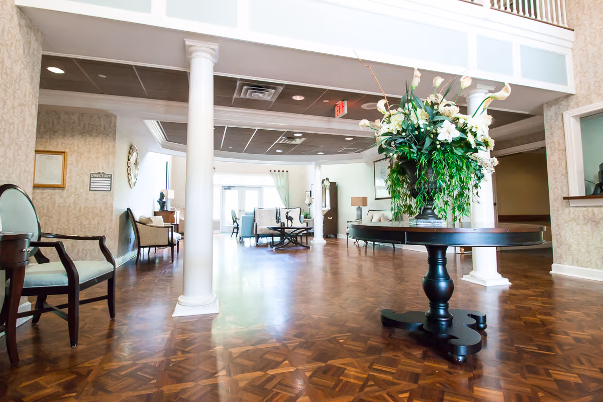 Spacious senior living facility lobby with polished wood floors, white columns, a central round table topped with a large floral arrangement, and seating areas in the background.